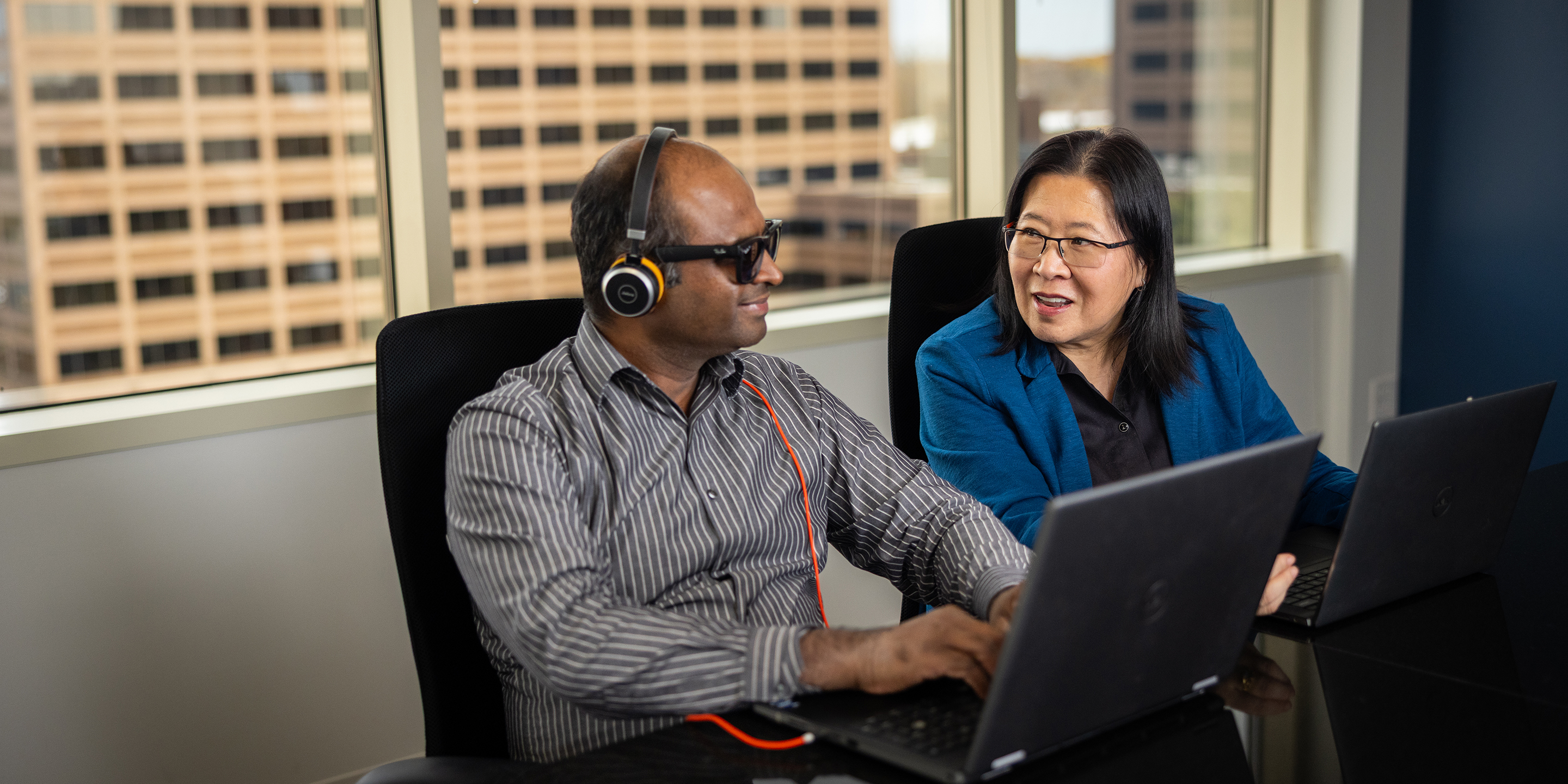 Two professionals working on laptops in a meeting room with large windows and office buildings visible in the background.