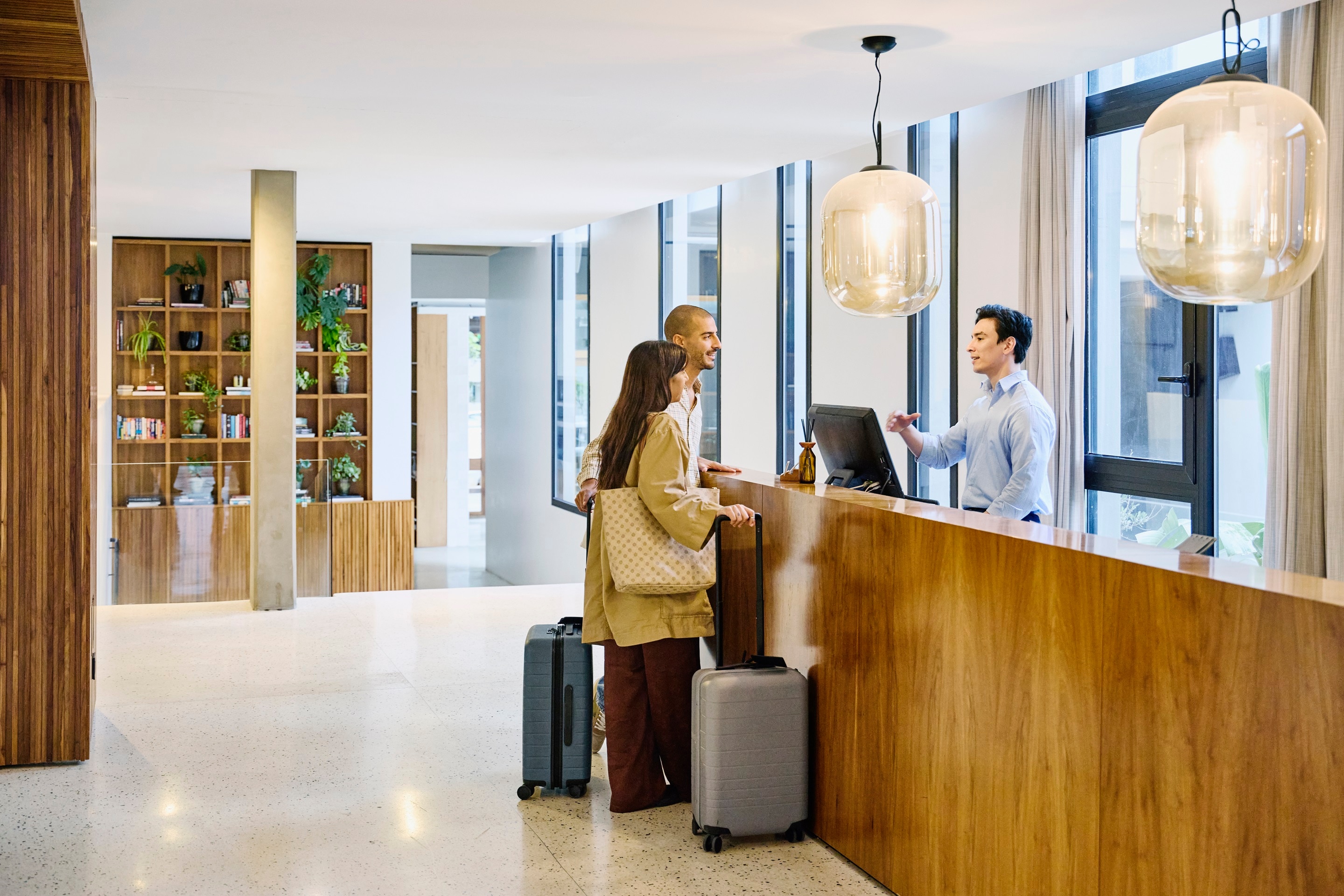 Wide shot of male hotel receptionist checking in guests with suitcases at hotel lobby front desk