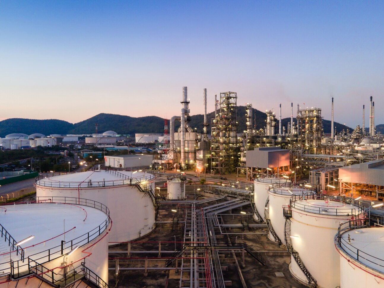 Industrial refinery complex with cylindrical tanks, pipes, and illuminated towers under an evening sky.